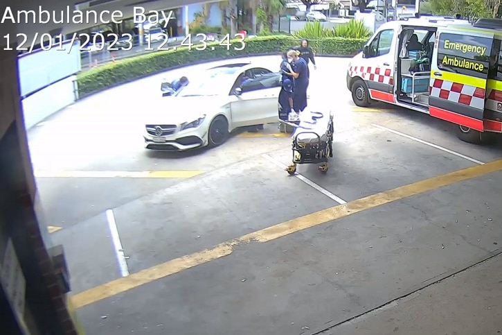 Paramedics in uniform lean into a white car while a man walks around it