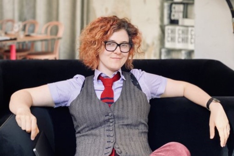 A woman with curly red hair and glasses poses for a photo on a black sofa wearing a grey pinstripe vest and maroon pants