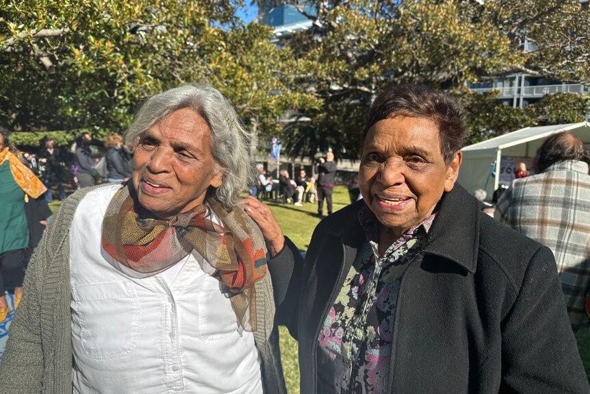 Two older Aboriginal women smile at an outside event in a green park.