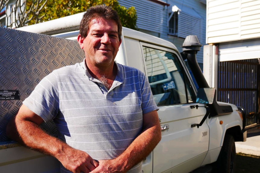 50 ish tradie smirks bemusedly at the camera while leaning up against his ute