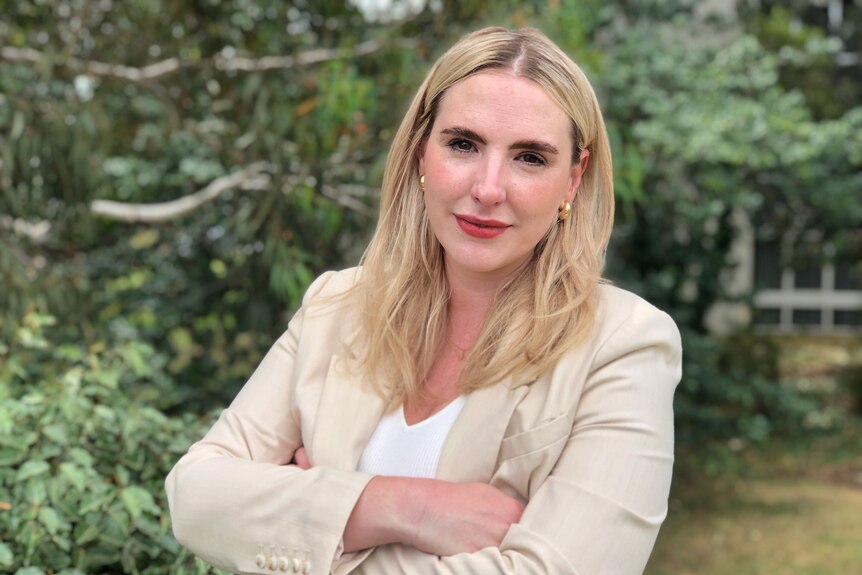 A woman with blond hair wearing a beige blazer stands with her arms crossed