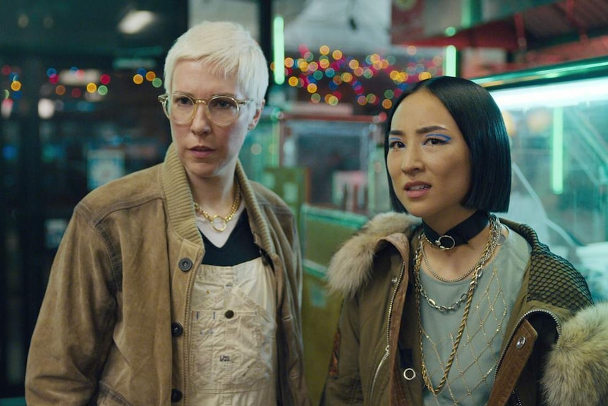 2 women in bodega, 1 has short platinum blonde hair & round clear-rimmed glasses, other has straight black bob & blue eyeshadow.