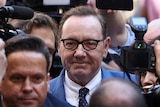 Actor Kevin Spacey, wearing glasses and a blue suit, is surrounded by a press pack.