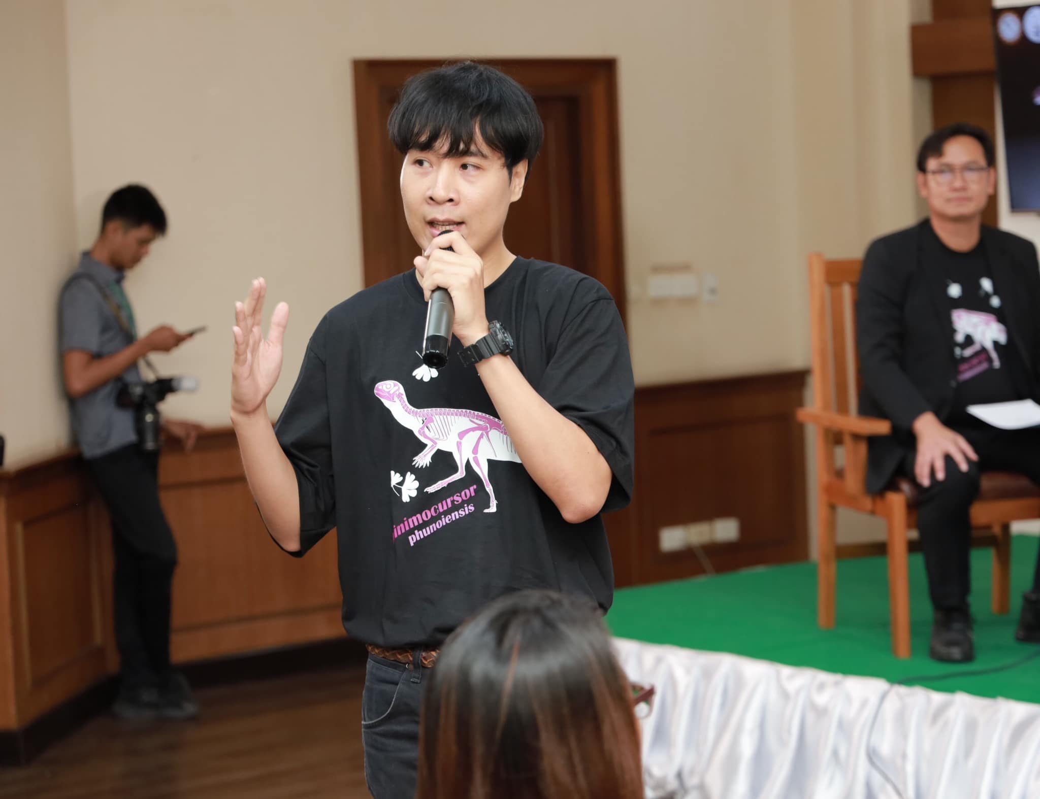 Sita Manitkoon speaks into a microphone, wearing a tshirt with a Minimocursor phunoiensis skeleton on it