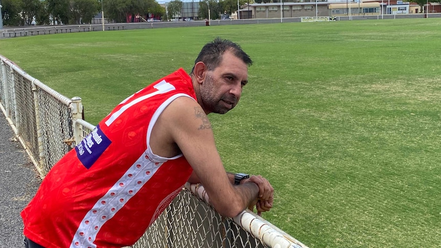 a man wearing a red football guernsey leans on the fence of a country aussie rules oval