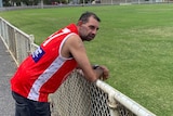 a man wearing a red football guernsey leans on the fence of a country aussie rules oval