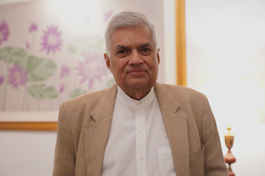 A man with white hair and wearing a tan blazer smiles at the camera.