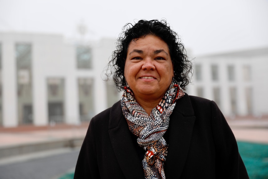 An Indigenous woman with short curly hair smiles into the camera with a government building behind her