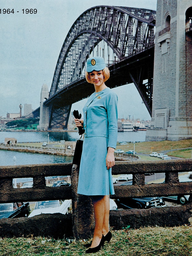 An old photo shows a woman in a light blue dress and hat standing in front of a view of the Sydney Harbour Bridge.