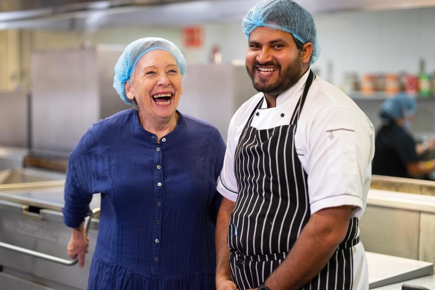 A woman in a blue shirt and blue hair net stands smiling next to a chef in a white shirt, apron and hair net.