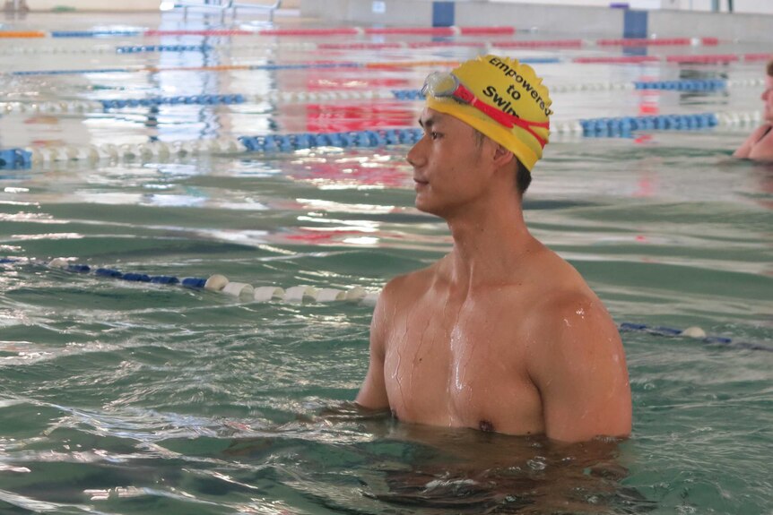 A young Asian man in yellow swimming cap looks to the side as he stands in indoor pool in complex.