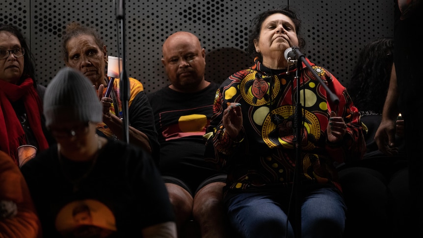 A woman claps her hands while listening to music, surrounded by mourners holding candles. 