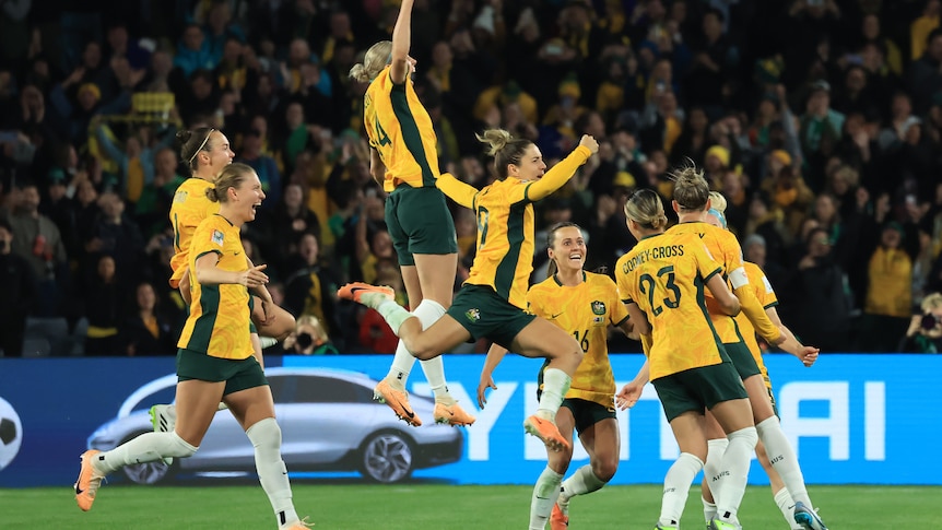 Australian female soccer players jump high in celebration in front of a crowd of fans