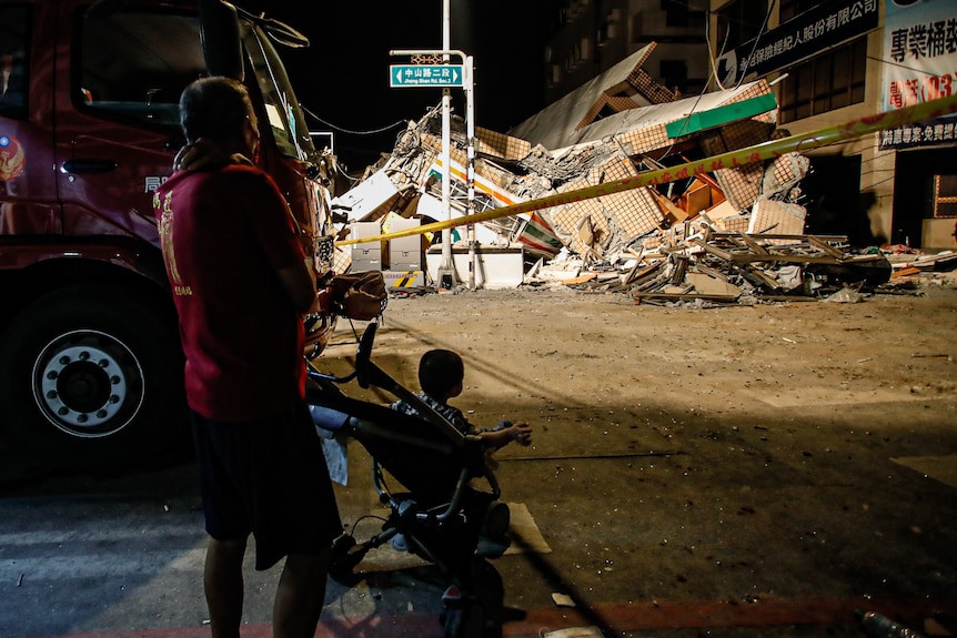 A person with a child in a pram looks at a collapsed building blocked by yellow tape. 