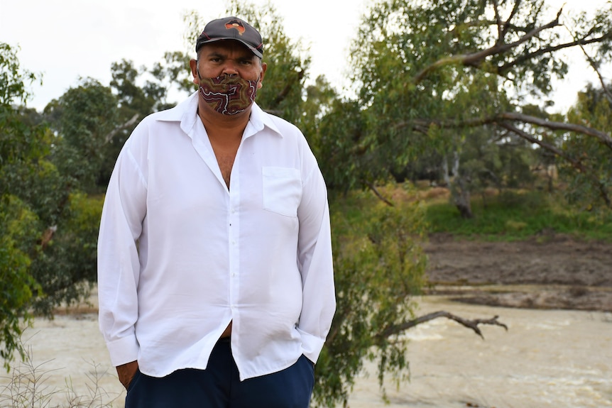 A man with a white shirt and cap stands in front of a river.