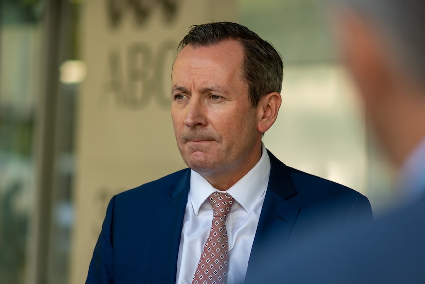 A middle aged man wearing blue suit, white shirt and patterned tie standing in front of light yellow wall.