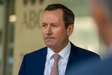 A middle aged man wearing blue suit, white shirt and patterned tie standing in front of light yellow wall.