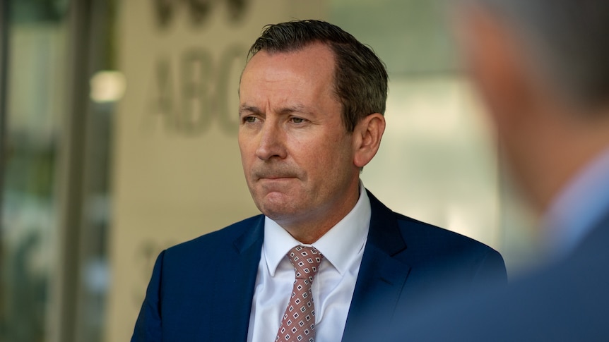 A middle aged man wearing blue suit, white shirt and patterned tie standing in front of light yellow wall.