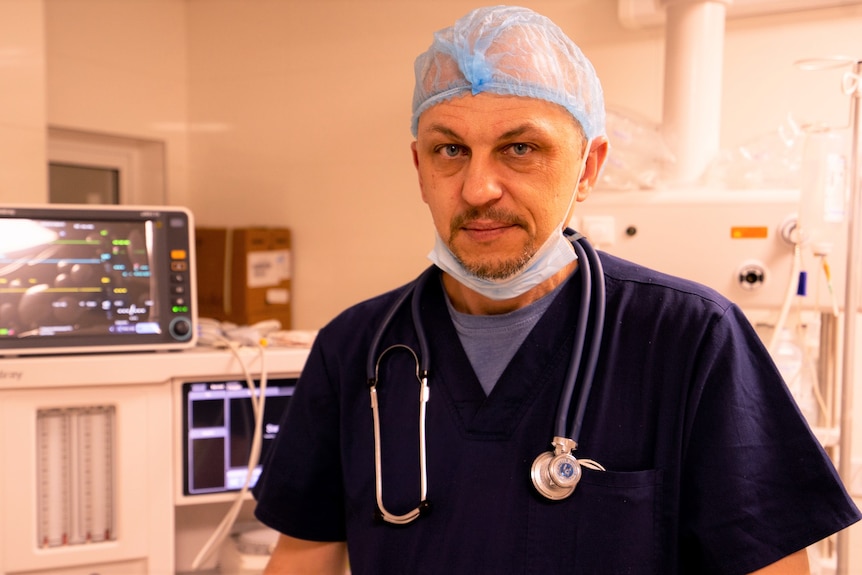 A man dressed in blue scrubs and wearing a hairnet and stethoscope smiles.