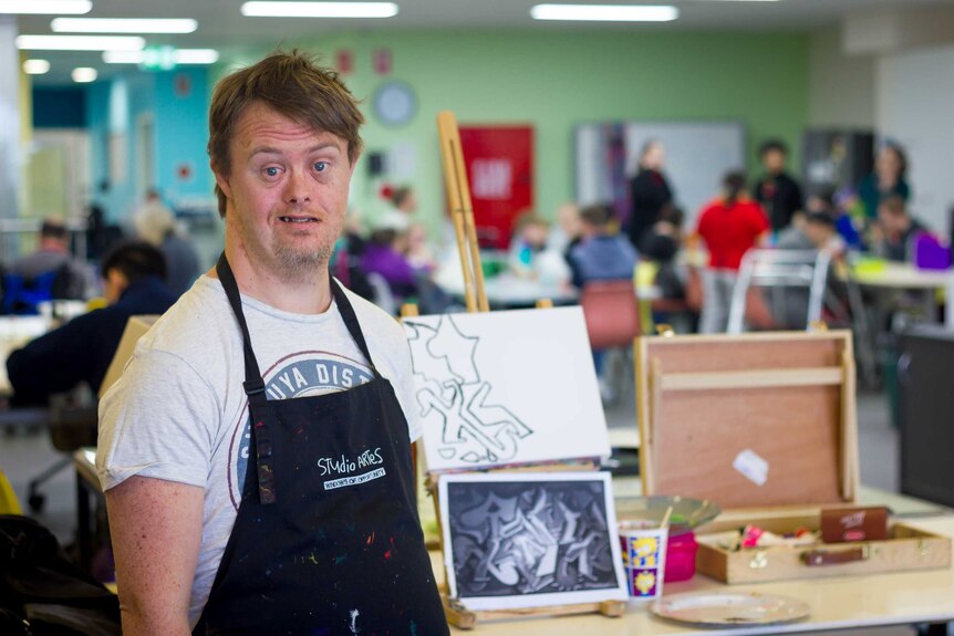 A 33-year-old man with Down syndrome stands in front of artworks at an art class.