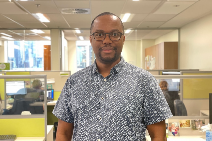 A man with short hair and a short beard and glasses standing in an office.