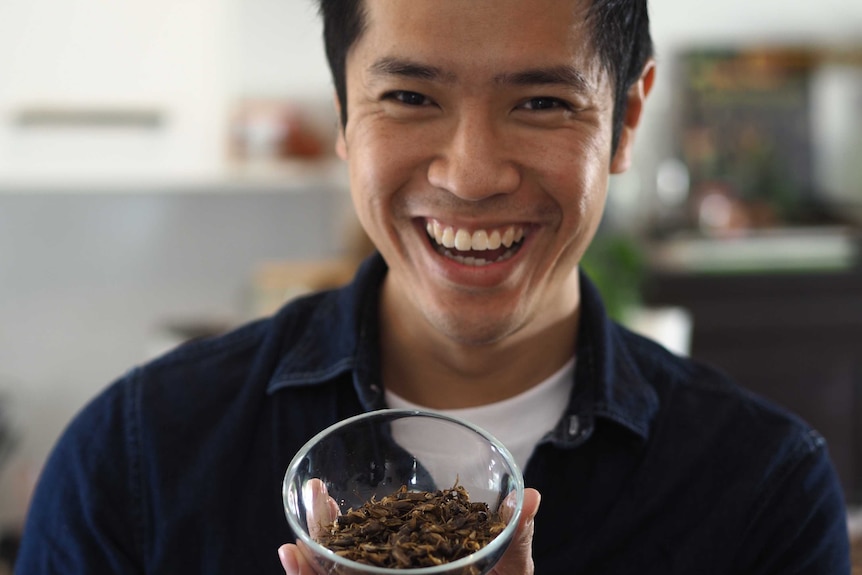 A grinning man holds a bowl of dried grasshoppers.