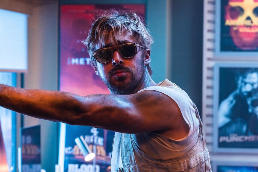 Ryan Gosling in aviator sunglasses and a sleeveless vest leans an arm against a doorframe