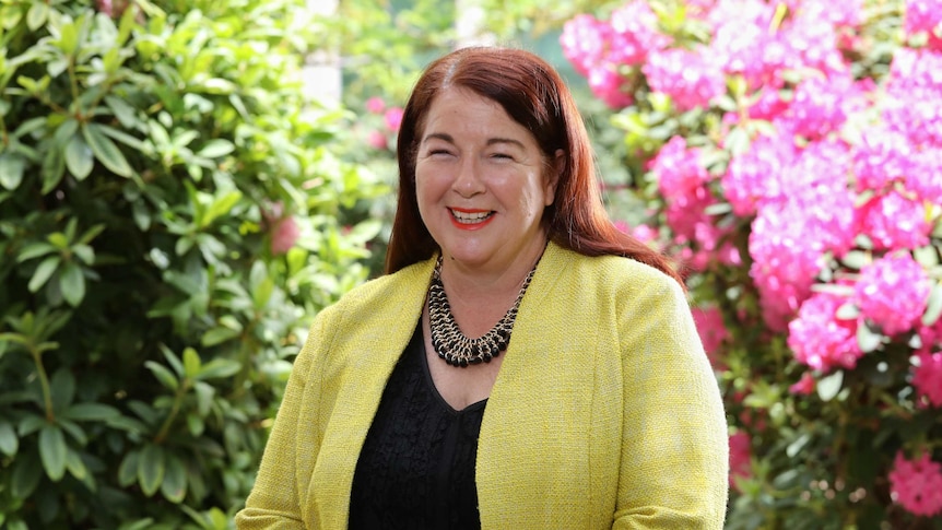 Melissa Price smiles at the camera. Behind her is a garden with pink flowers. She is wearing yellow