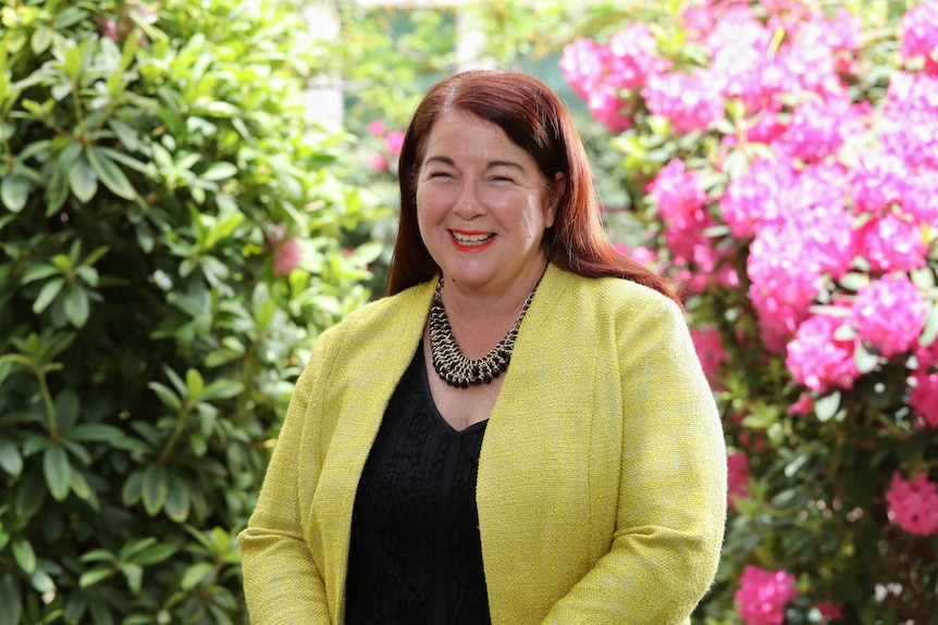 Melissa Price smiles at the camera. Behind her is a garden with pink flowers. She is wearing yellow