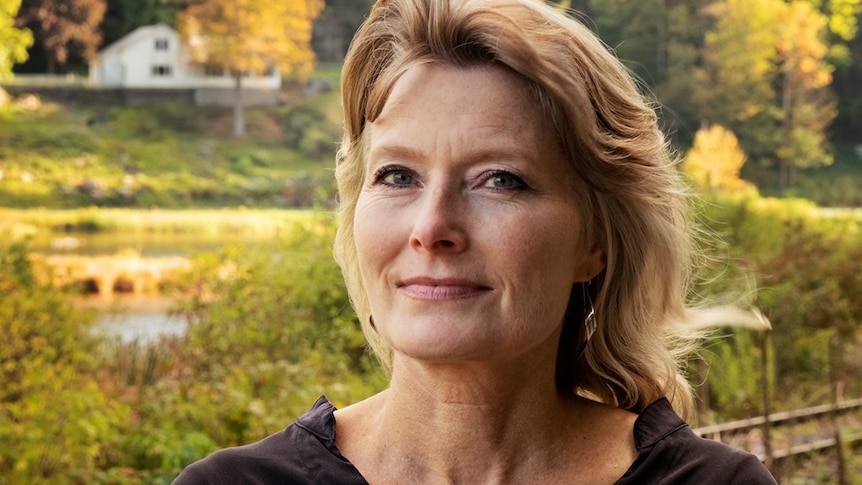 A middle-aged white woman with brown eyes, blonde hair and a slight smile stands in front of a verdant riverbank