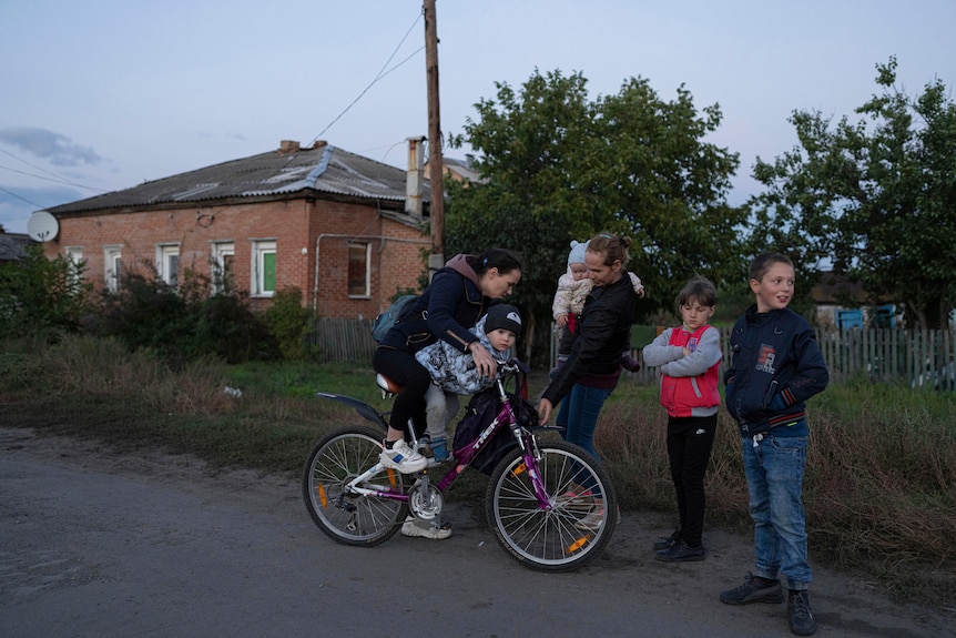 A woman and child on a bike stop on a road next to another woman holding a baby and two children. 