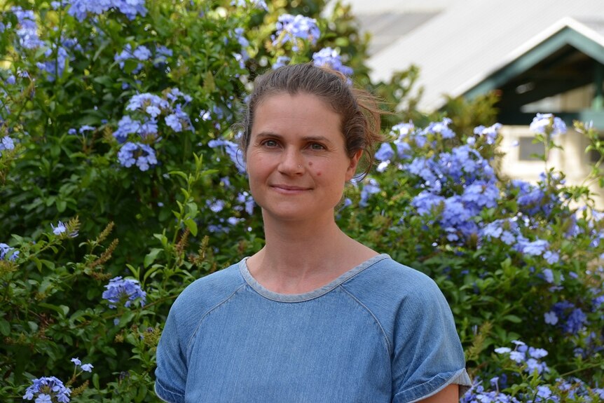 A smiling woman with brown hair tied back, blue tee, in front of flowering bush with blue flowers.