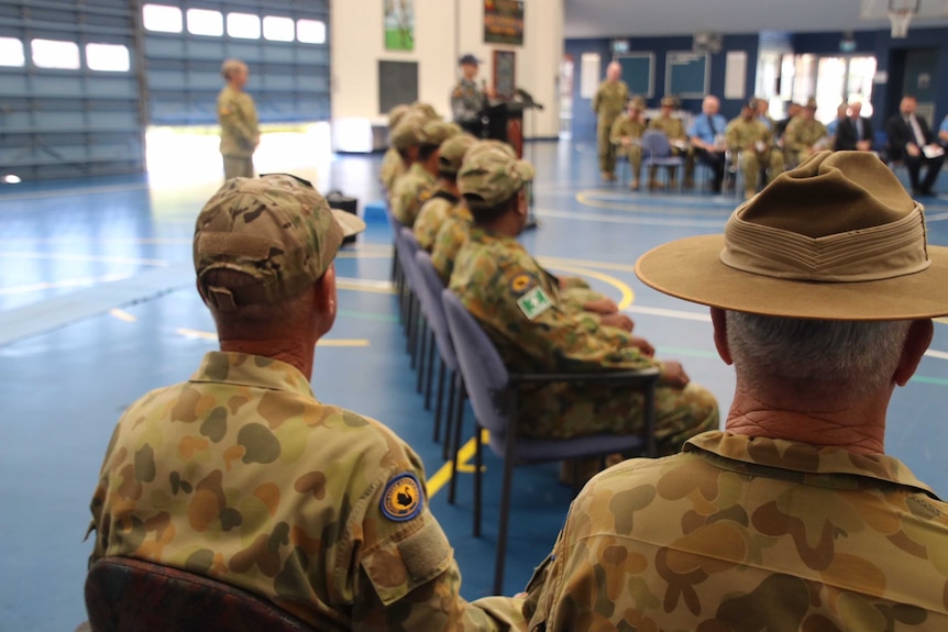 Two men dressed in military uniform look towards a group of teenage boys in camouflage uniforms sitting in a line.