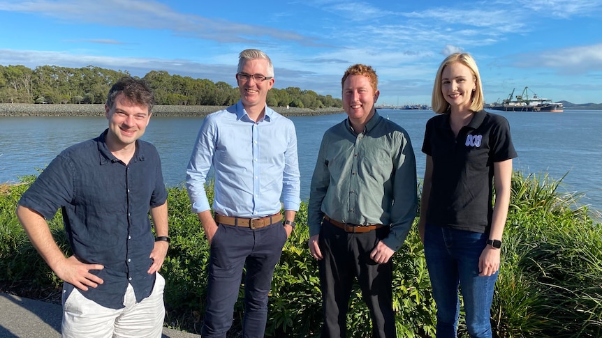 Three men and one woman smiling to camera with blue water bay and ship in background.