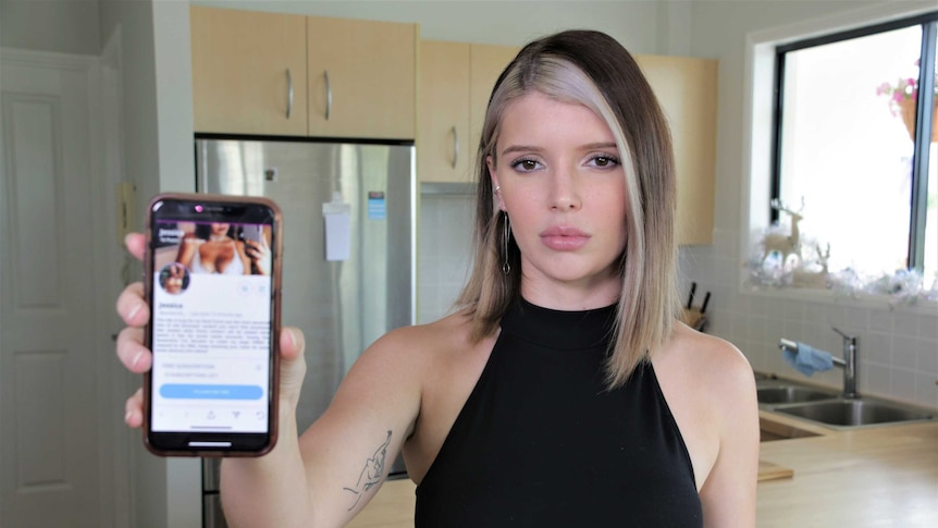 Woman in her 20s holding a mobile phone to the camera with a profile page on the screen. She does not look impressed.