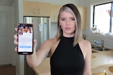 Woman in her 20s holding a mobile phone to the camera with a profile page on the screen. She does not look impressed.