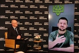 A suited man seated on a stage chats with a man who is shown on a large video screen.