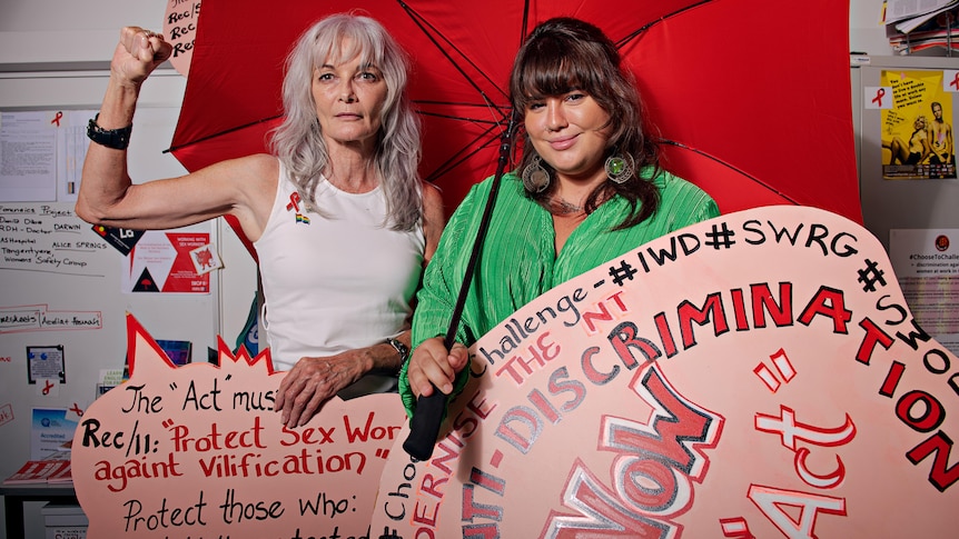 Two women stand holding a red umbrella and red and pink placards in an office. 