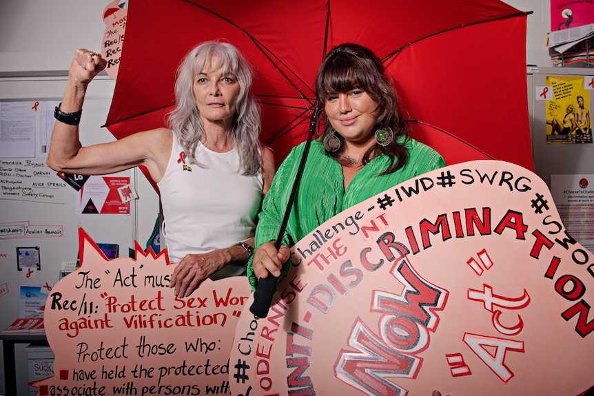 Two women stand holding a red umbrella and red and pink placards in an office. 