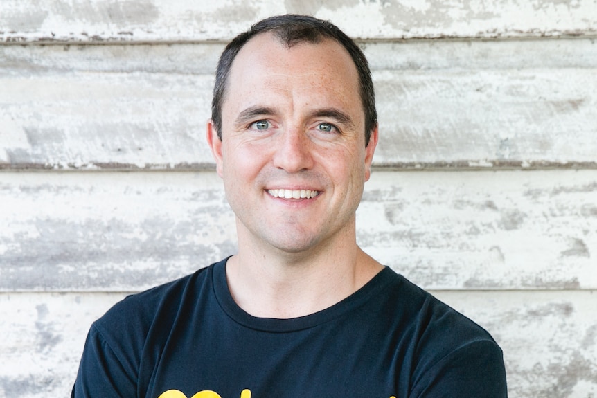 A dark-haired man in a black and yellow shirt standing against a rustic wood house wall and he is smiling