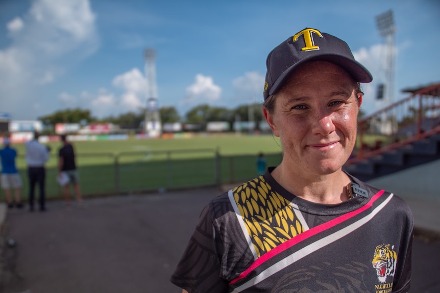 A woman wearing club colours stands and smiles at the camera in front of a footy field