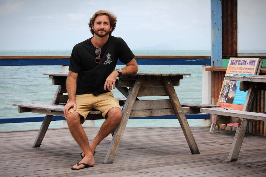 A casually dressed man sits at an empty table on a deck overlooking the ocean