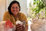 A women smiles as she holds different varieties of mushrooms, with bags of mushrooms in front of her on a table. 