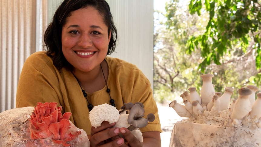 A women smiles as she holds different varieties of mushrooms, with bags of mushrooms in front of her on a table. 