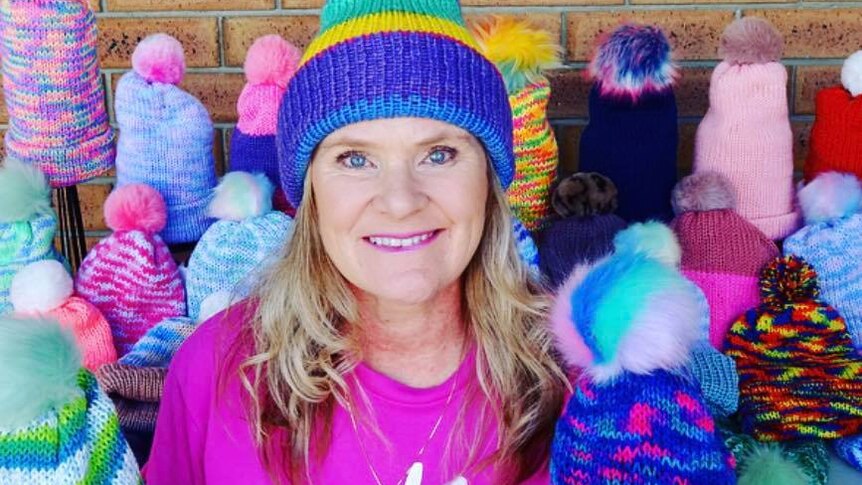 smiling lady in colourful pom-pom hat and bright pink t-shirt surrounded by knitted hats