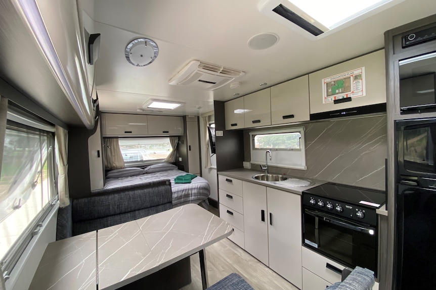 The interior of a caravan shows a small kitchen table, bed, and kitchen area.