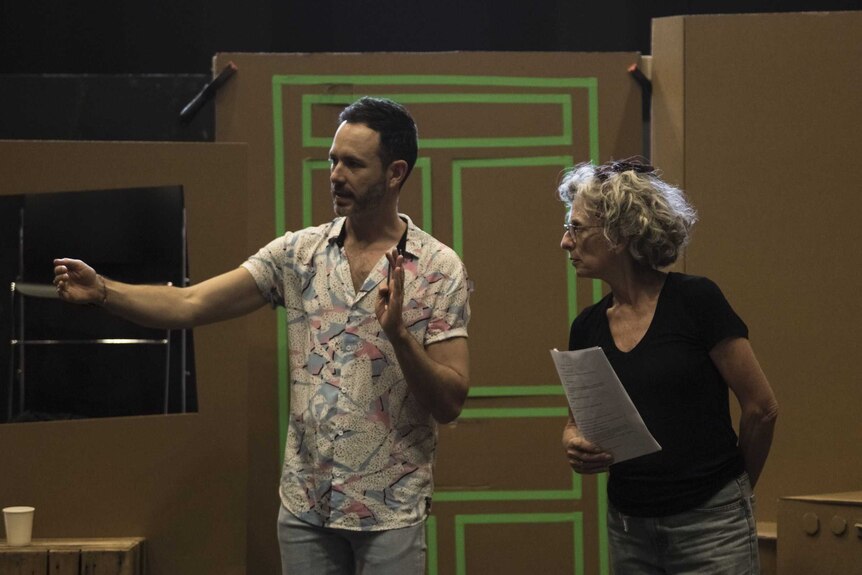 Artistic director Luke Kerridge and a woman with a script in hand on a stage set with cardboard boxes that make a house