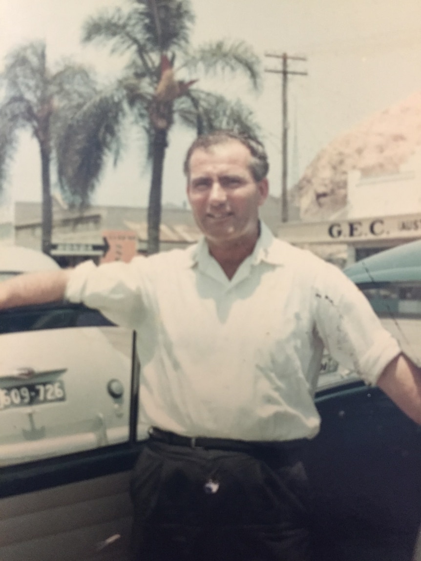 A man stands in front of a car in a photo taken in the 1960s.