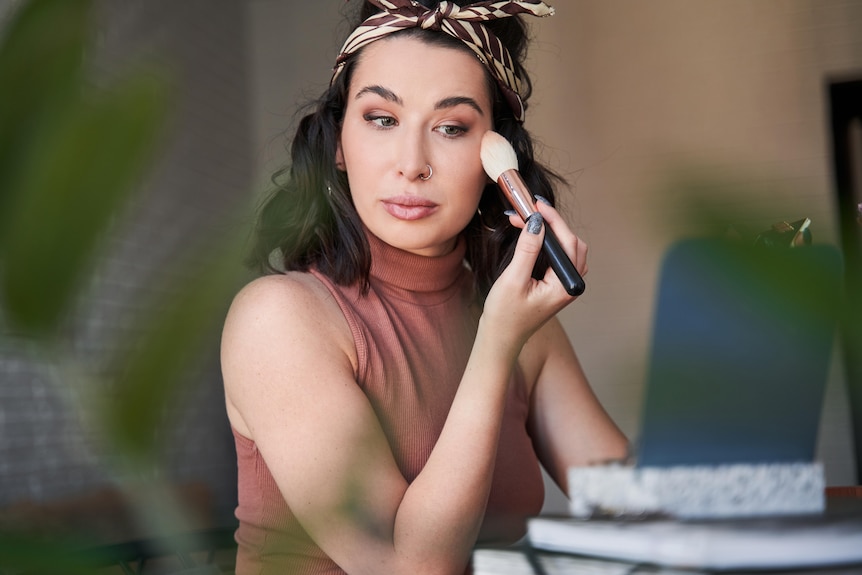 Young woman doing her makeup in front of the mirror. She holds a fluffy makeup brush and has a headband in her hair.