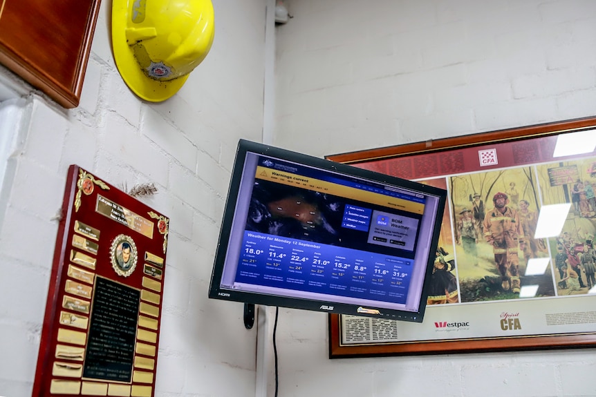A TV screen on a wall shows weather forecasts, CFA helmet and photos also on the wall. 